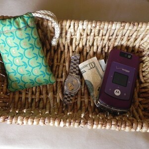 May include: A woven wicker basket with a green fabric pouch, a silver wristwatch, a purple cell phone, and a $10 bill.