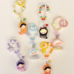 Könnte beinhalten: Sammlung bunter Schlüsselanhänger mit kleinen Figuren in verschiedenen Outfits, darunter ein Bär, ein Pilz und ein Teufel. Jeder Schlüsselanhänger hat ein Perlenband mit passendem Farbschema und einen Metallverschluss. Die Schlüsselanhänger befinden sich auf weißem Hintergrund.