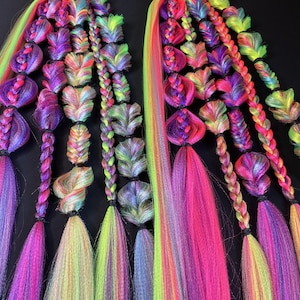 May include: Two sets of colorful hair extensions with a variety of braided and twisted designs. The extensions feature vibrant hues of pink, purple, yellow, and green, creating a rainbow effect. The hair is styled in various shapes.