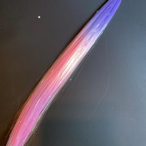 May include: A vibrant hair extension with a gradient of colors, transitioning from pink to purple. The extension is secured with a black elastic band and features shimmering strands for added sparkle. The hair piece is designed to add length and color to any hairstyle.