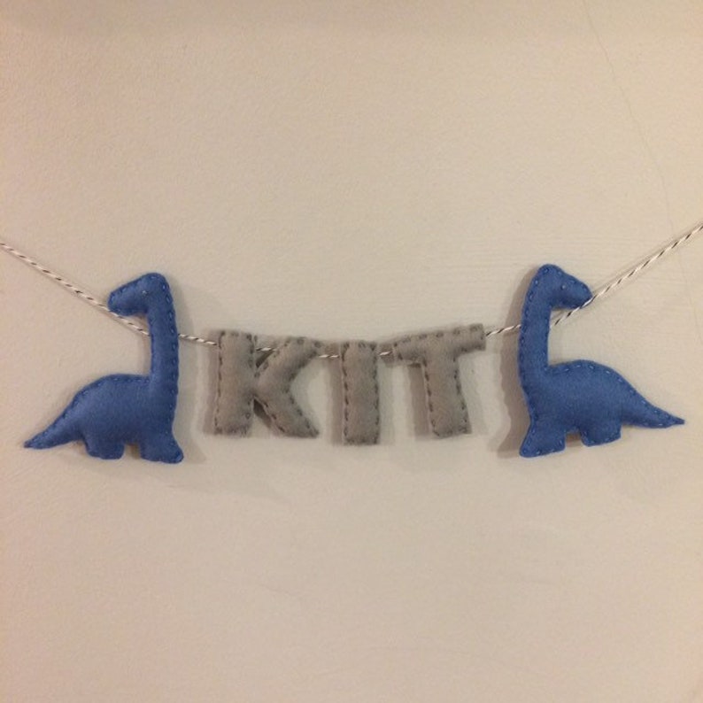 May include: A felt banner with the name "KIT" in gray letters, flanked by two blue dinosaur figures. The banner is strung on a white and silver cord, suitable for nursery or children's room decor.