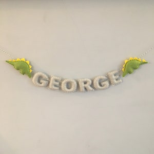 May include: A felt banner spelling "GEORGE" in light gray letters, strung on a white and gray twine. The banner is decorated with two green dinosaur shapes with yellow spines, one on each end. A decorative item for a child's room.