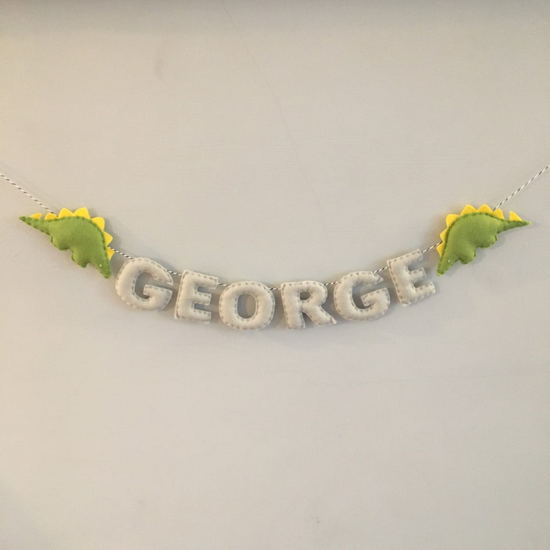 May include: A felt banner spelling "GEORGE" in light gray letters, strung on a white and gray twine. The banner is decorated with two green dinosaur shapes with yellow spines, one on each end. A decorative item for a child's room.