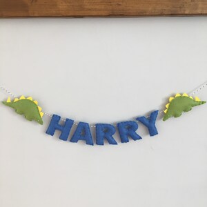 May include: A felt banner with the name "HARRY" in blue letters, flanked by green dinosaur shapes with yellow spines. The banner is strung on a white cord and hangs against a white wall.