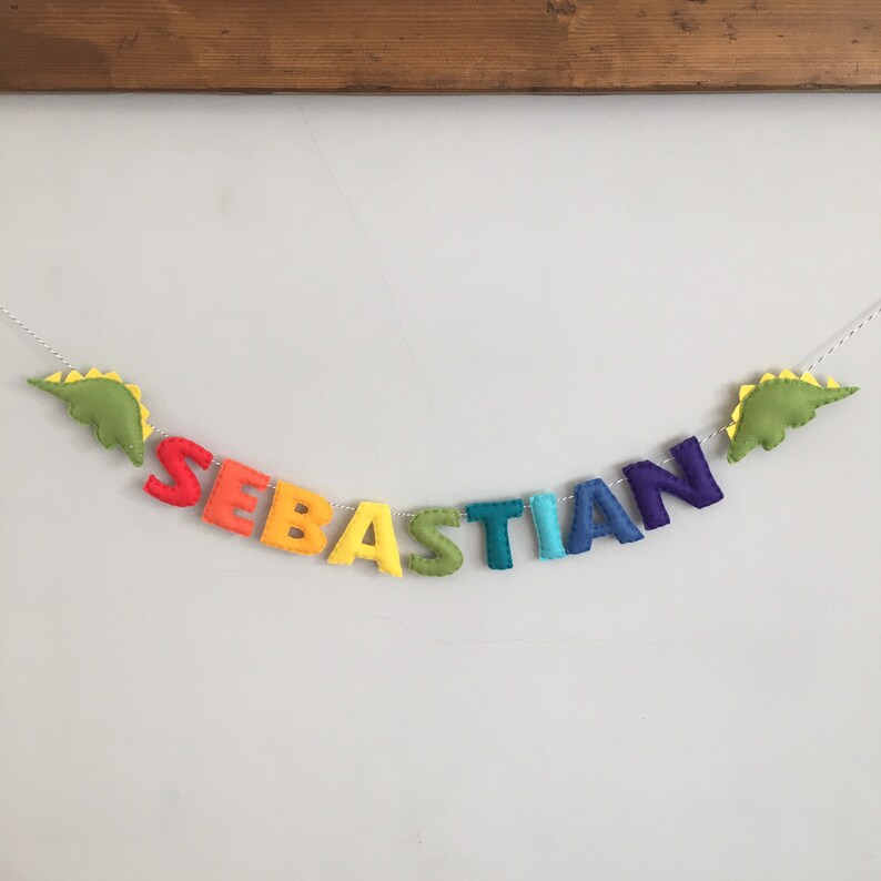 May include: A colorful felt banner spelling "SEBASTIAN" in rainbow hues. The letters are strung on a white cord, with green dinosaur shapes at each end. The banner is a decorative item.