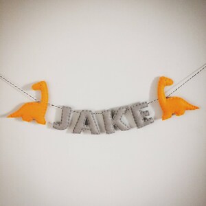 May include: A felt banner with the name "JAKE" in gray letters, stitched with white thread. Two orange dinosaur figures flank the name. The banner hangs against a white background.