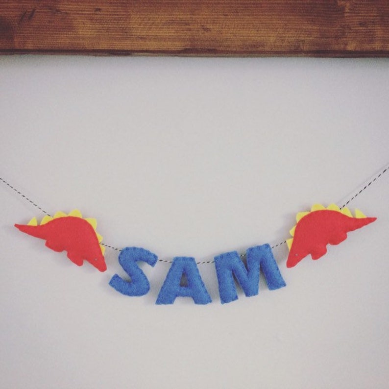 May include: A felt banner spelling "SAM" in blue letters, with red and yellow dinosaur accents on a string. The banner is hanging against a white wall, with a wooden frame at the top.