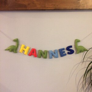 May include: A felt banner spelling "HANNES" in colorful letters, with a green dinosaur on each end. The letters are yellow, red, green, light blue, and dark blue. The banner is strung on a white cord.