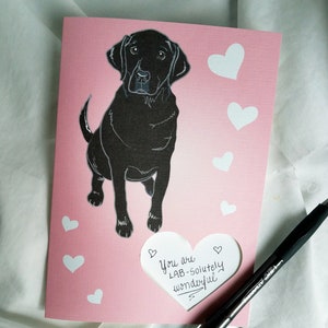 May include: A pink card with a black Labrador Retriever illustration and white hearts. The card has a cut-out heart shape with the text "You are LAB-solutely wonderful" written inside.