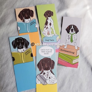 May include: Four bookmarks featuring a German Shorthaired Pointer dog with different book-themed illustrations and text. The bookmarks are on a gray surface. The text on the bookmarks includes "Perfect Pointers", "How to Play Fetch", "Dog Tails", "Let's curl up with a good book!" and "Birdwatching".