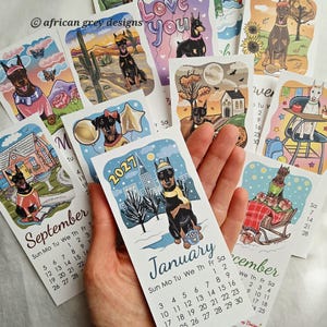 May include: A collection of illustrated 2027 calendars featuring Doberman Pinschers in various seasonal settings. Each calendar page displays a month with dates and a unique dog-themed illustration, with the "January" calendar page in focus.
