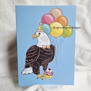 May include: A greeting card featuring an illustrated bald eagle wearing a party hat and bow tie, holding balloons and a cupcake. The eagle is brown, white, and yellow, with a light blue background. The balloons are pink, tan, orange, yellow, and green.