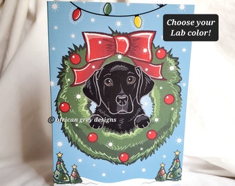May include: A black Labrador Retriever puppy peeking out of a Christmas wreath with red berries and a red bow. The wreath is decorated with twinkling lights and snowflakes. The background is a blue sky with snow falling. The text "Choose your Lab color!" is in the upper right corner.