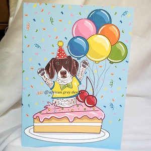 May include: A birthday greeting card featuring a cartoon dog wearing a party hat and a yellow shirt, emerging from a slice of pink frosted cake. Colorful balloons and confetti decorate the light blue background. The card is on a white surface.