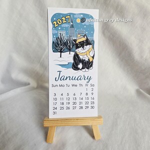 K&ouml;nnte beinhalten: Ein Januar 2027 Kalender mit einer schwarz-wei&szlig;en Katze mit gelbem Hut und Schal, vor einer verschneiten Stadtlandschaft. Der Kalender wird auf einer kleinen Holzstaffelei pr&auml;sentiert.