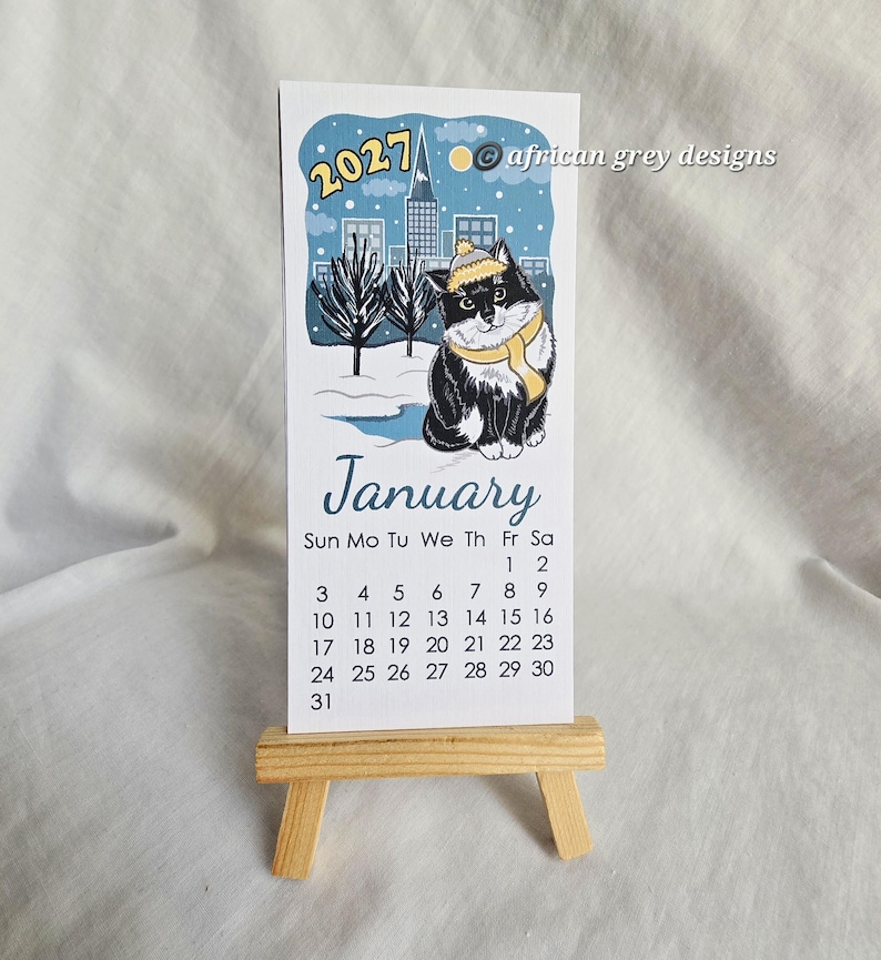 K&ouml;nnte beinhalten: Ein Januar 2027 Kalender mit einer schwarz-wei&szlig;en Katze mit gelbem Hut und Schal, vor einer verschneiten Stadtlandschaft. Der Kalender wird auf einer kleinen Holzstaffelei pr&auml;sentiert.