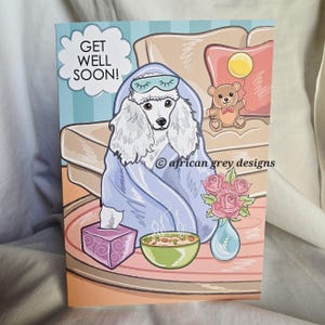 Könnte beinhalten: Eine Grußkarte mit einer Cartoon-Illustration eines weißen Pudels, der eine Schlafmaske trägt und in eine blaue Decke gehüllt ist. Die Karte trägt die Aufschrift "GET WELL SOON!" und zeigt eine Schüssel Suppe, eine Taschentuchbox und eine Vase mit Rosen.