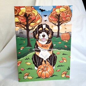 May include: A Bernese Mountain Dog puppy wearing a black and orange bandana with the word "BOO!" on it, sitting in a green grassy field with fall leaves and a pumpkin. The dog is looking at the camera with its tongue sticking out. There are two trees in the background with orange, yellow, and red leaves.