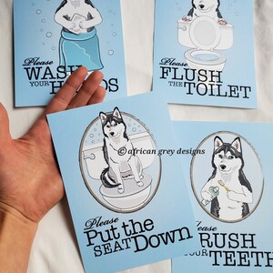 May include: Four blue signs with a cartoon husky dog on each. The signs have instructions for bathroom etiquette. The first sign says "Please Wash Your Hands". The second sign says "Please Flush The Toilet". The third sign says "Please Put The Seat Down". The fourth sign says "Please Brush Your Teeth".
