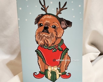 May include: A Christmas card featuring a cartoon Brussels Griffon dog wearing a red and green Santa suit with reindeer antlers. The dog is holding a wrapped gift and is standing on a light blue background with white snowflakes.