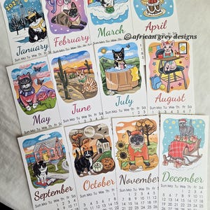 K&ouml;nnte beinhalten: Ein 2027-Kalender mit monatlichen Illustrationen von Katzen in verschiedenen Umgebungen. Jeder Monat zeigt eine einzigartige Szene mit Katzenmotiven, mit Daten und Wochentagen. Der Kalender enth&auml;lt den Text "African Grey Designs".
