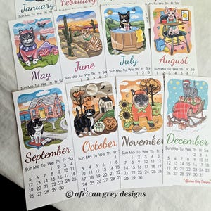 K&ouml;nnte beinhalten: Eine Reihe von illustrierten Monatskalendern mit skurrilen Katzenillustrationen. Jeder Monat zeigt eine andere Katze in einer saisonalen Umgebung, mit dem Monatsnamen und den Daten darunter. Der Kalender wurde von African Grey Designs entworfen.