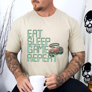 以下が含まれることがあります： 緑色の「Eat Sleep Game Repeat」の文字とレトロなビデオゲームコントローラーのグラフィックが描かれたベージュのTシャツ。