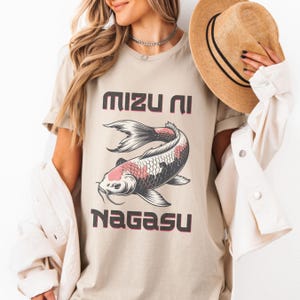 Peut inclure: T-shirt beige avec une illustration de carpe koï et le texte japonais "Mizu Ni Nagasu" en rose foncé. La carpe koï présente des détails noirs, blancs et roses. Le t-shirt a des manches courtes et un col rond.