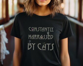 Puede incluir: Una camiseta negra con el texto "CONSTANTLY HARASSED BY CATS" en una fuente estilizada. El texto está rodeado de pequeñas imágenes de gatos.