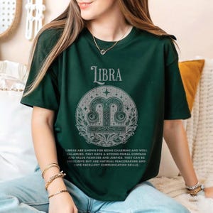 Könnte beinhalten: Dunkelgrünes T-Shirt mit dem Sternzeichen Waage. Das Design zeigt das Wort "LIBRA" über einem kreisförmigen Symbol. Text darunter beschreibt die Eigenschaften der Waage. Ein bequemes, lässiges T-Shirt.