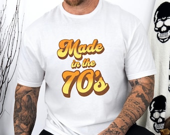Puede incluir: Camiseta blanca con un gráfico de estilo retro que dice "Made in the 70's" en naranja y amarillo.
