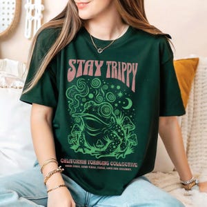 Może przedstawiać: Ciemnozielona koszulka z grafiką palącej żaby, grzybów i różowym napisem "STAY TRIPPY". Poniżej widnieje napis "CALIFORNIA FORAGING COLLECTIVE" oraz "GOOD VIBES, PEACE, LOVE AND HARMONY."