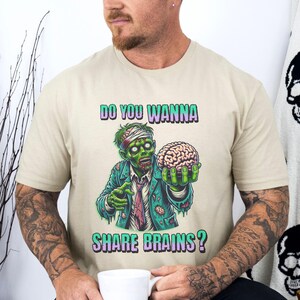 Puede incluir: Una camiseta beige con un zombie verde sosteniendo un cerebro y el texto "Do you wanna share brains?"