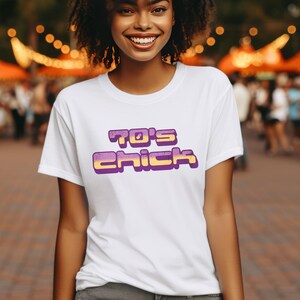 以下が含まれることがあります： レトロなフォントで「70's Chick」と書かれた、紫と黄色のグラフィックが施された白いTシャツです。