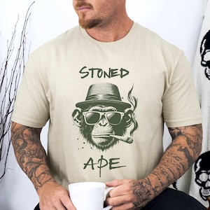 May include: A beige t-shirt with a green illustration of a monkey wearing sunglasses and a hat, smoking a cigarette. The text "STONED APE" is printed on the shirt.