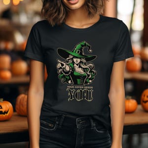 Op de afbeelding: Zwart T-shirt met een groene heks-afbeelding die een puntige hoed draagt en haar hand uitsteekt. De tekst "Your Coven Needs You" is onder de heks gedrukt.