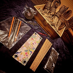 May include: A collection of tools and accessories, including rope bundles in a wooden box, scissors, and a decorative fabric piece with a peacock design. Other items include wooden sticks and metal tools, all arranged on a dark purple surface.