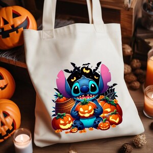 May include: A cream-coloured canvas tote bag featuring a colourful graphic of a blue alien character holding a glowing jack-o'-lantern, surrounded by several other pumpkins, bats, and a spider web, with two sturdy shoulder straps at the top.