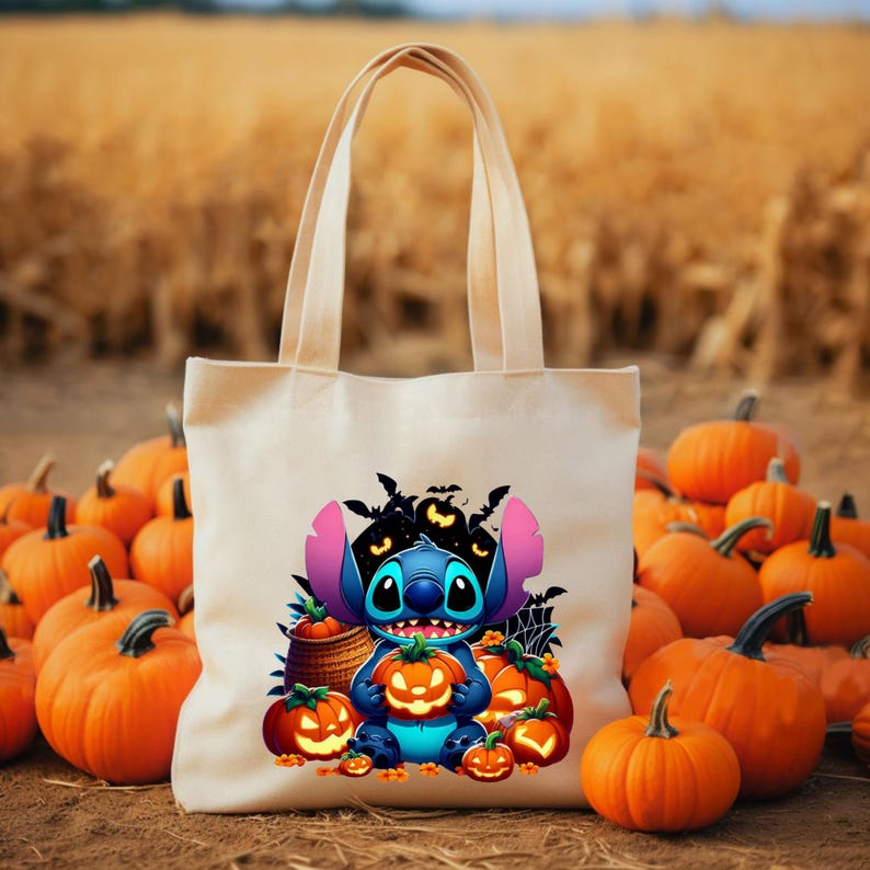 May include: A beige canvas tote bag featuring a colourful graphic of a blue alien character holding a glowing orange jack-o'-lantern, surrounded by smaller pumpkins, a woven basket, a spider web, and black bats against a dark background.
