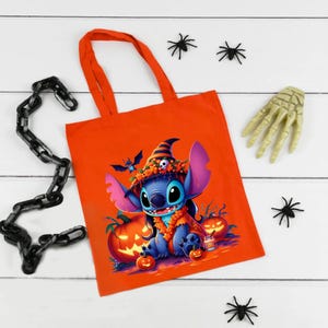 May include: An orange tote bag featuring a graphic of a blue alien character wearing a witch hat and orange floral garland, surrounded by jack-o'-lanterns. The design includes a small skull on the hat and a tiny bat flying above the character.
