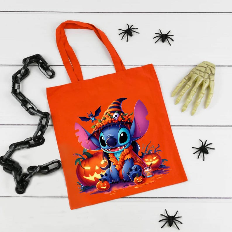 May include: An orange tote bag featuring a graphic of a blue alien character wearing a witch hat and orange floral garland, surrounded by jack-o'-lanterns. The design includes a small skull on the hat and a tiny bat flying above the character.