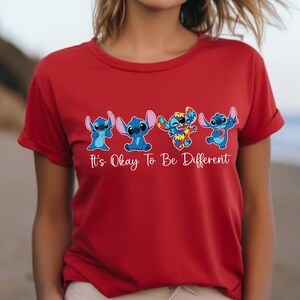 May include: Red t-shirt featuring four blue cartoon characters with the text "It's Okay To Be Different". The characters are in different poses, with one character having a colorful, puzzle-like design.