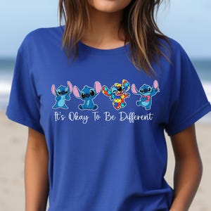 May include: Royal blue t-shirt featuring four cartoon characters in various poses. The characters are primarily blue with pink accents. The text "It's Okay To Be Different" is printed in white below the characters.