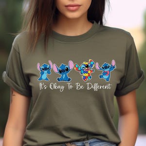 May include: Olive green t-shirt featuring four cartoon Stitch characters in various poses. One Stitch is multi-colored, and the text "It's Okay To Be Different" is printed below the characters. The shirt has short sleeves.