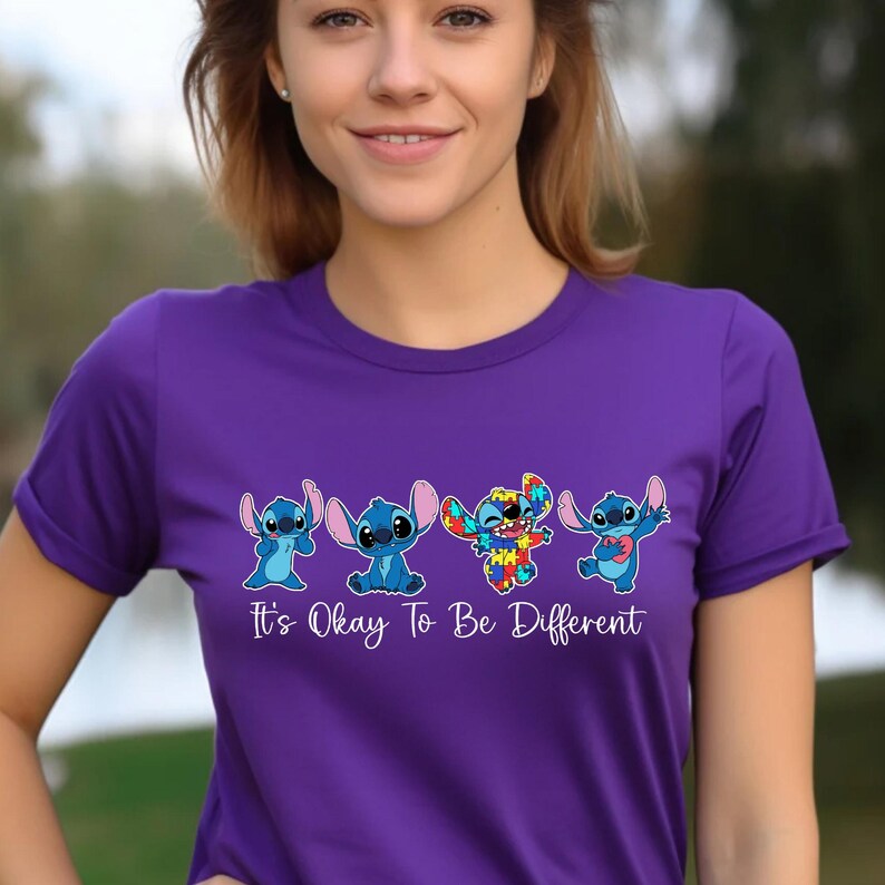 May include: A purple t-shirt featuring four cartoon Stitch characters in various poses. The Stitch characters are in shades of blue and rainbow colors. The text "It's Okay To Be Different" is printed in white below the characters.