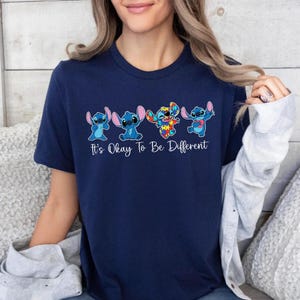 May include: Navy blue t-shirt featuring four cartoon characters in different poses. The characters are blue with large ears. One character is designed with a colorful puzzle pattern. The text "It's Okay To Be Different" is printed below the characters.