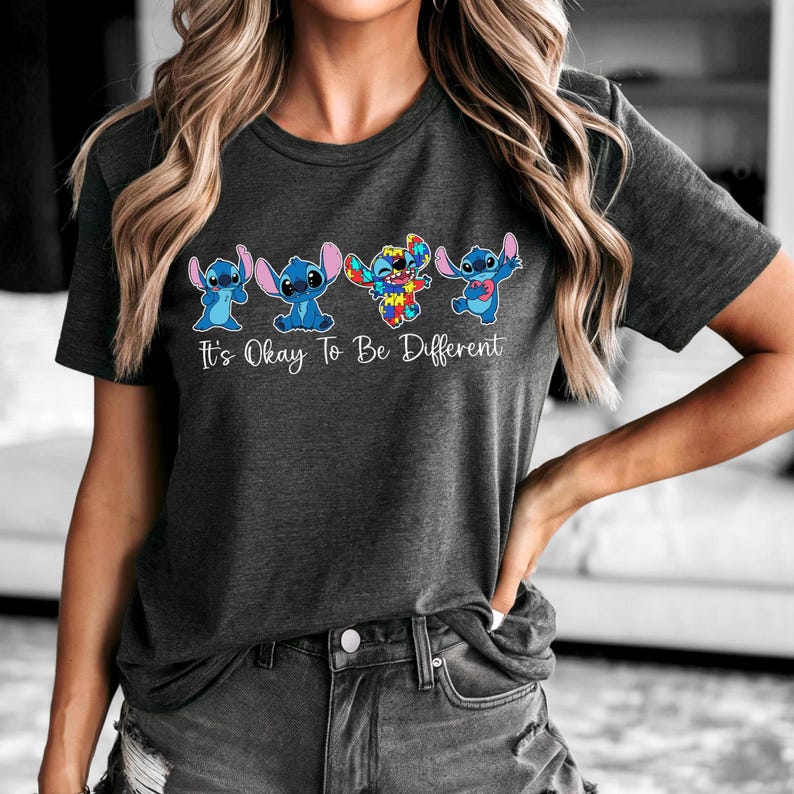 May include: Dark gray t-shirt featuring four cartoon Stitch characters in blue and rainbow colors. The text "It's Okay To Be Different" is printed below the characters in white. The shirt is a casual wear item.