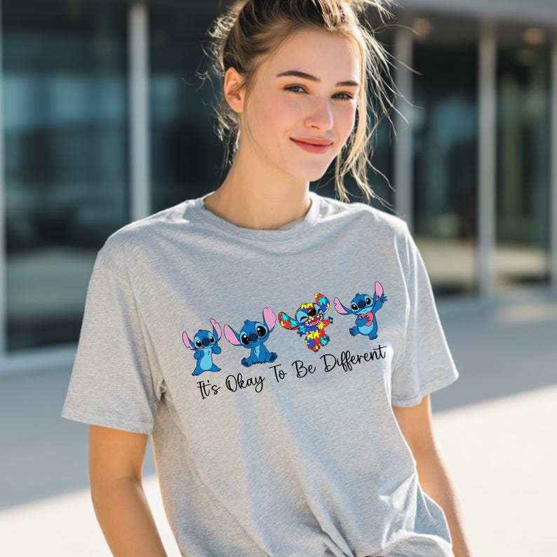 May include: A light gray t-shirt featuring four cartoon Stitch characters in various poses. One Stitch is multi-colored. The text "It's Okay To Be Different" is below the characters. The shirt is made of a soft material.