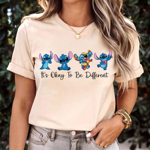 May include: Beige t-shirt featuring four cartoon Stitch characters in various poses. One Stitch is designed with a colorful patchwork pattern. The text "It's Okay To Be Different" is printed below the characters in black.