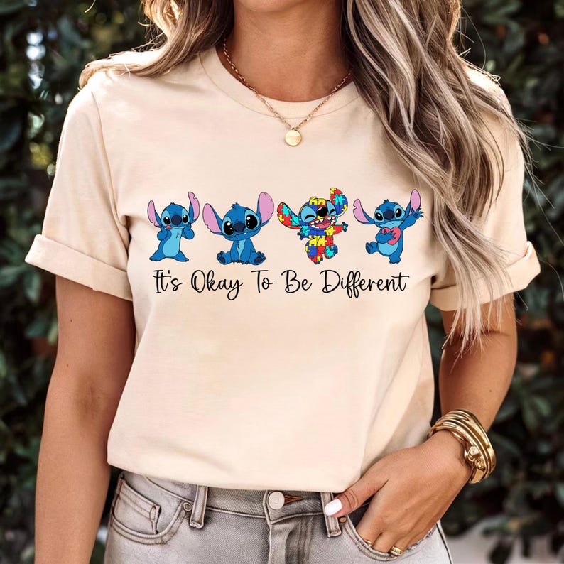 May include: Beige t-shirt featuring four cartoon Stitch characters in various poses. One Stitch is designed with a colorful patchwork pattern. The text "It's Okay To Be Different" is printed below the characters in black.